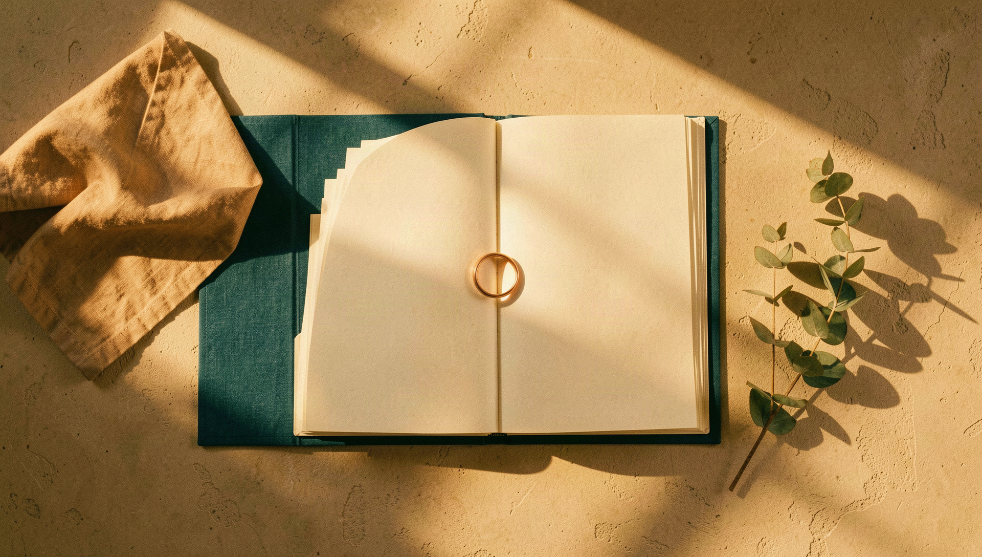 Editorial still life of a teal cloth folder with blank pages fanned open, a copper ring resting on the fold, linen and eucalyptus beside it in golden light